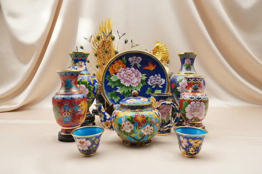 An exquisite collection of handcrafted cloisonné enamel art, including a teapot, teacups, and decorative vases adorned with vibrant peony floral motifs and gold filigree, displayed on a beige silk background.